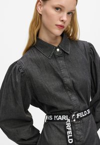 Ciemnoniebieska koszula dżinsowa z kołnierzykiem, bufiastymi rękawami i czarno-białym, markowym paskiem. Posiada centralny guzik i fakturowane wykończenie.