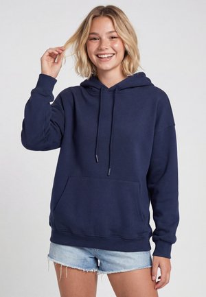 Tânără zâmbitoare cu păr blond scurt, purtând un hanorac albastru închis și pantaloni scurți din denim bleu deschis, atingându-și părul cu o mână.