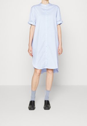 Lichtblauwe blousejurk met korte pofmouwen, ronde hals en een gebogen zoom. Gecombineerd met grijze geribbelde sokken en zwarte instapschoenen.