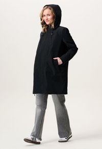 Vrouw in een zwarte gewatteerde jas met capuchon, met handen in de zakken, grijze jeans en witte sneakers met zwarte strepen, staande op een effen lichte achtergrond.