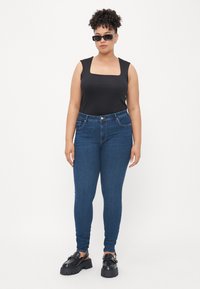 Black square-neck tank top, blue slim-fit jeans, and chunky black shoes. The outfit features a smooth texture and fitted design.