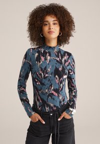 Top à manches longues bleu avec des motifs abstraits rose et noir, col haut, design ajusté, associé à un jean foncé.