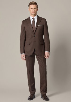 Mężczyzna stojący w dopasowanym brązowym garniturze z białą koszulą, brązowym krawatem, poszetką i ciemnobrązowymi eleganckimi butami na beżowym tle.