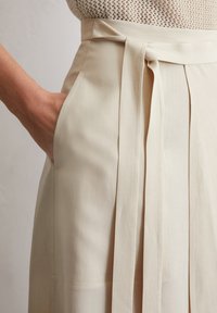 Light beige wrap skirt with a tied waist, smooth texture, side pockets, and clean stitching. Captures drape and tailored design details.