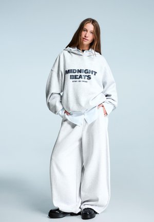 Sivá oversized mikina s nápisom "MIDNIGHT BEATS", v kombinácii so širokými sivými teplákmi a čiernymi platformovými topánkami. Hladká textúra, oblečenie v neformálnom štýle.