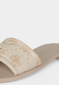 Μπεζ σανδάλι τύπου slip-on με υφασμάτινο λουράκι με ανοιχτά λουλουδάτα σχέδια και μεταλλικό τρίγωνο με το λογότυπο Guess στην κορυφή.