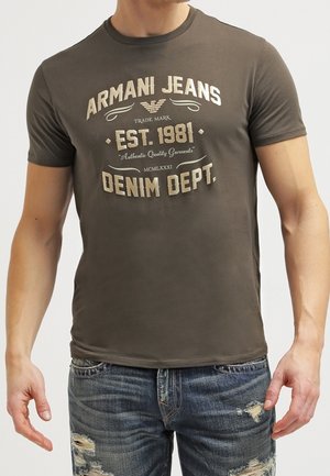 Man die een nauwsluitend donkergroen Armani Jeans T-shirt met bedrukte tekst en versleten blauwe spijkerbroek draagt.
