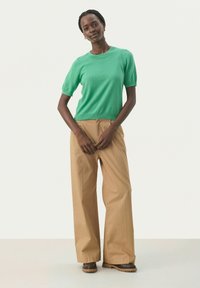 Grön kortärmad stickad topp med rundad hals, i kombination med högmidjade, vida beige byxor. Modellen står framför en neutral bakgrund.