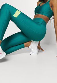 Set di reggiseno sportivo e leggings di colore teal, realizzato in materiale elasticizzato. I leggings presentano una patta di accento verde e dettagli perforati sulla coscia.