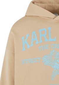 Felpa con cappuccio beige caratterizzata da testo blu "KARL THE ORIGINAL STREET" con una grafica di un uccello stilizzato, realizzata in tessuto morbido.
