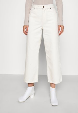 Frau trägt hoch taillierte, offwhite weite Hose, beige geripptes Langarmoberteil und weiße Knöchelstiefel mit Blockabsätzen, steht auf grauem Boden.