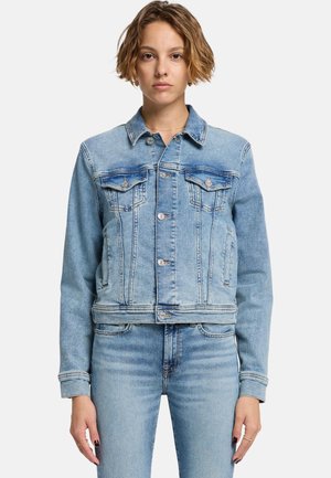 Kvinde med kort bølget hår iført en lys blå denimjakke og matchende denimjeans, stående mod en ensfarvet hvid baggrund.