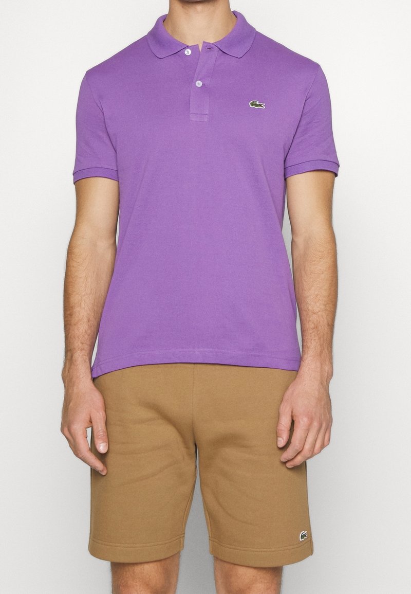 Homme portant un polo à manches courtes violet et un short marron, debout devant un fond blanc uni.