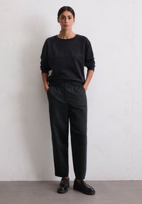 Une femme aux cheveux noirs en queue de cheval basse porte un sweat-shirt noir Marc O'Polo, un pantalon noir et des mocassins noirs, se tenant contre un mur blanc.