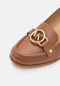 Mocassin en cuir marron avec une boucle circulaire dorée portant les initiales "MK" sur la partie avant supérieure de la chaussure.