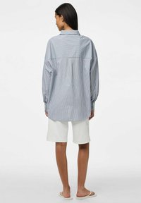 Prugasta košulja dugih rukava u plavoj i bijeloj boji, opuštenog kroja, s detaljem na ramenu. Uparena s bijelim kratkim hlačama i ravnim sandalama.