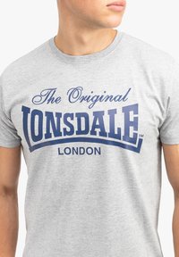 Pilka medvilninė marškinėlių su trumpomis rankovėmis, kuriuose mėlyna spalva užrašytas tekstas "The Original LONSDALE LONDON" išlenktoje formoje. Lygus paviršius, laisvas kirpimas.