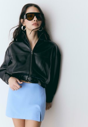 Kobieta w za dużej czarnej skórzanej kurtce, niebieskiej mini spódnicy z rozcięciem, dużych kolczykach kołach i przyciemnianych okularach przeciwsłonecznych na białym tle.