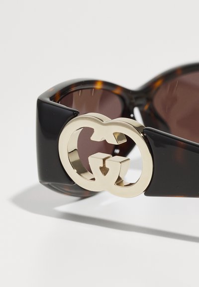 Juodos ir kilpinės didelės saulės akiniai su ryškiu auksiniu tarpusavyje susikertančių GG logotipu ant šventyklų ir rudi objektyvai.