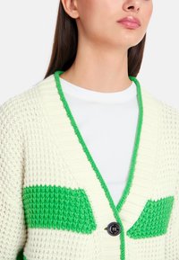 Strickjacke in Creme mit grünen Streifen, V-Ausschnitt, geripptem Kragen und einer einzigen Knopfleiste. Texturiertes Material über die gesamte Fläche.