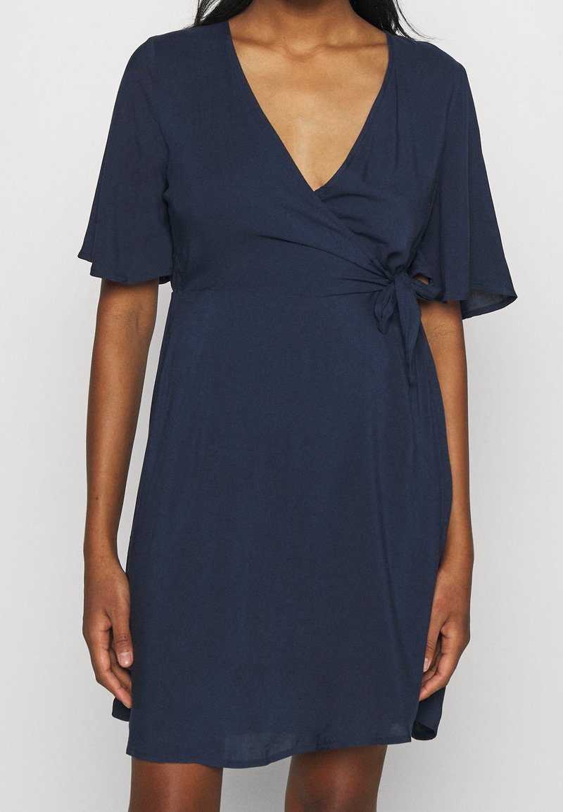 Femme portant une robe portefeuille bleu marine avec des manches à volants et un nœud sur le côté, debout devant un fond clair uni.