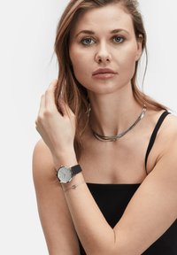 Sølvfarvet ur med en rund urskive og sort rem; modellen bærer en reflekterende, buet metalhalskæde og et delikat armbånd.