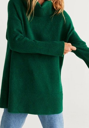 Femme tirant sur la manche d'un pull à col roulé vert surdimensionné, portant un jean bleu clair, sur un fond clair uni.