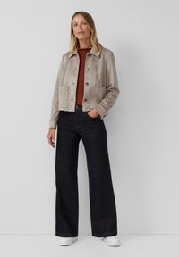 Une femme portant une veste beige boutonnée, un haut texturé rouille, un jean large foncé et des baskets blanches, se tenant devant un fond clair.