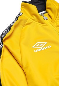 Geel ritsjack van glad polyester, met zwarte zijaccenten met een tribale patroon en een witte "UMBRO"-logo op de borst.