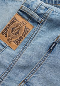 Jeans de mezclilla de color azul claro con un parche de cuero marrón con una marca en relieve, costuras visibles y una pequeña etiqueta negra en el lateral.