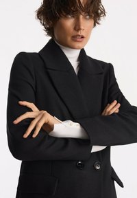 Czarny płaszcz dwurzędowy o fakturowanym wykończeniu, z szerokimi klapami i guzikami. Biała golfowa bluzka pod spodem z detalami w postaci złotych guzików.