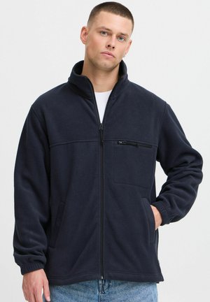 Veste en polaire marine avec col montant, fermeture éclair sur le devant, poche poitrine zippée et deux poches latérales. Texture douce et poignets côtelés.