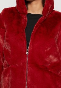 Rød faux fur jakke med hætte, med en lynlås foran, blød tekstur og rynkede detaljer på siderne. Sort skjorte under.