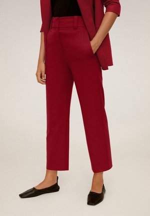 Pantaloni a sigaretta rossi, accorcati, indossati con un blazer abbinato e ballerine nere, con una mano in tasca, su uno sfondo neutro.