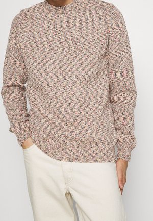 Mann trägt einen mehrfarbigen Strickpullover mit langen Ärmeln und cremefarbenen Hosen, die Hände teilweise in den Taschen, vor einem schlichten Hintergrund.