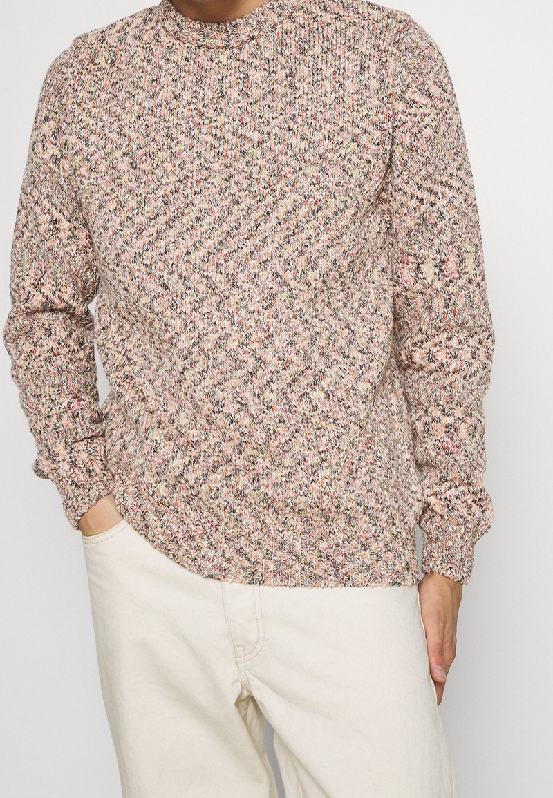 Homme portant un pull en maille multicolore à manches longues et un pantalon crème, les mains partiellement dans les poches, sur un fond uni.