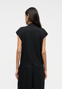 Femeie cu păr lung și închis la culoare, purtând un top negru fără mâneci și pantaloni negri, cu spatele spre privitor, pe un fundal simplu și deschis la culoare.