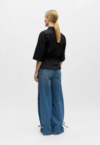 Femme à queue de cheval blonde portant un haut noir à manches courtes et un jean bleu pattes larges avec des liens au niveau de l'ourlet, vue de dos sur fond uni.