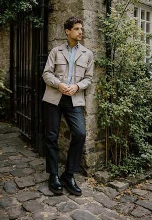 Mladić u bež jakni i crnim hlačama naslonjen na kameni zid kraj zelenila i željeznih vrata na kaldrmisanoj stazi.