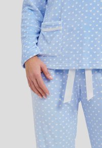 Pijamas de color azul claro con patrones de corazones blancos, que cuentan con un bolsillo en la parte superior y un lazo en los pantalones, hechos de tejido suave.