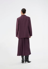 Γυναίκα με τα μαλλιά σε κότσο, που φοράει ένα μωβ μακρύ σακάκι και αντίστοιχη φούστα, με την πλάτη στραμμένη προς τον θεατή, συνδυασμένα με μαύρες ψηλές μπότες σε λευκό φόντο.