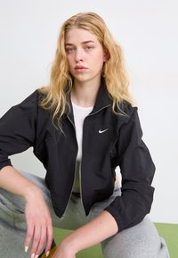 Juodas džemperis su užtrauktuku, pagamintas iš lengvos medžiagos, su elastingomis rankovėmis ir baltu Nike logotipu ant krūtinės, derinamas su pilkomis sportinėmis kelnėmis.