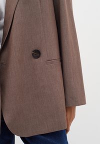 Blazer marrone a un bottone con rever indossato sopra una camicia bianca e jeans blu, vista parziale del torso e del braccio di una persona.