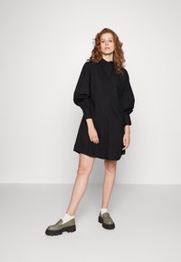 Vestido preto de botões com mangas bufantes, apresentando detalhes texturizados e um corte solto, combinado com sapatos chunky em tom de oliva e meias brancas.