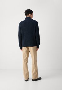 Suéter de canalé azul marino con cuello alto, de mangas largas y corte relajado, combinado con pantalones beige claros y zapatos negros.