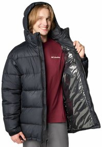 Giacca a vento nera con cappuccio, caratterizzata da sezioni trapuntate e un interno lucido con rifiniture argentate. Il modello indossa una maglietta color bordeaux sotto.