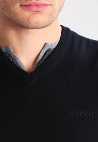 Marinblå stickad tröja med V-ringning, som har en grå inre krage och en diskret broderad logotyp på bröstet. Slät textur.