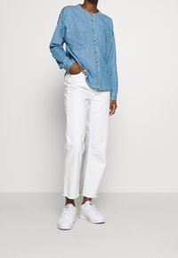Camisa de mezclilla azul clara con botones en el frente y dos bolsillos en el pecho, combinada con pantalones blancos de cintura alta y zapatillas blancas.