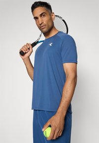 Blaustes Sport-T-Shirt mit kurzen Ärmeln und Rundhalsausschnitt; glatte Textur; Logo auf der Brust. Hält einen Tennisschläger mit einem Tennisball in der Hand.
