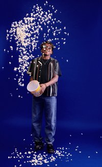 Osoba w pasiaku czarno-białym i niebieskich dżinsach trzyma żółty kubełek z popcornem, z którego popcorn się wysypuje, na niebieskim tle.